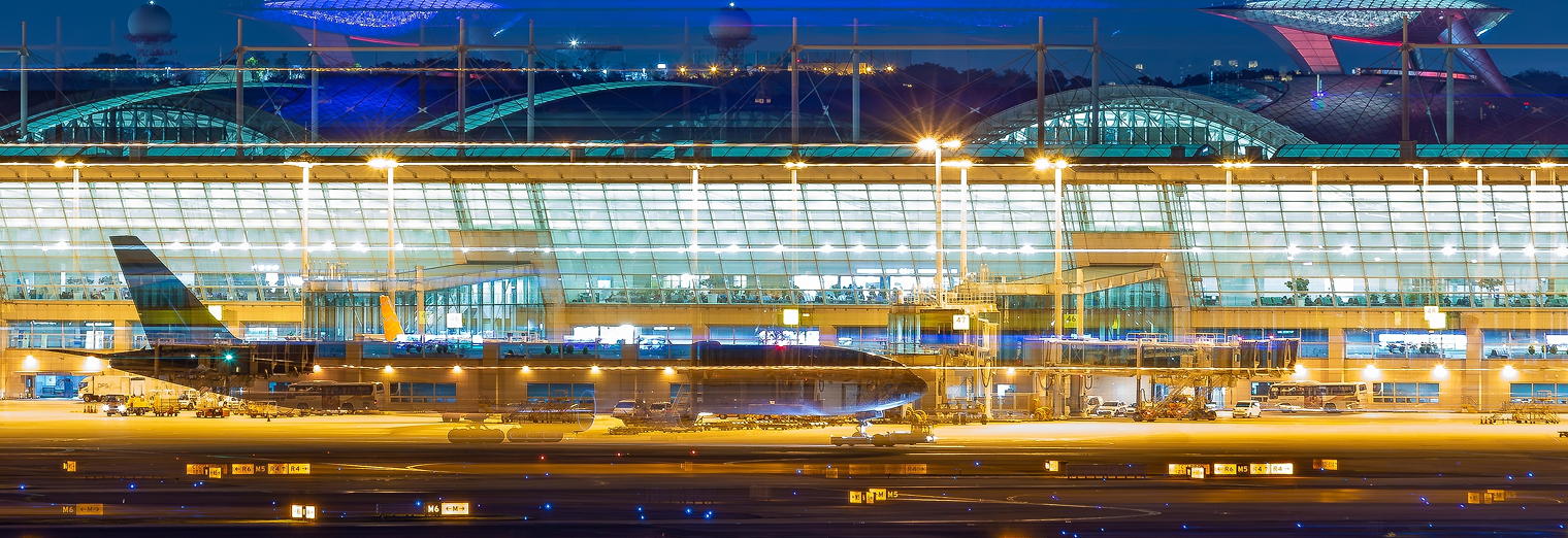 A passenger airplane parked on the tarmac at night in front of a brightly lit, modern airport terminal with large glass windows and a curved roof, conveniently located near WallyPark Airport Parking for easy travel access.