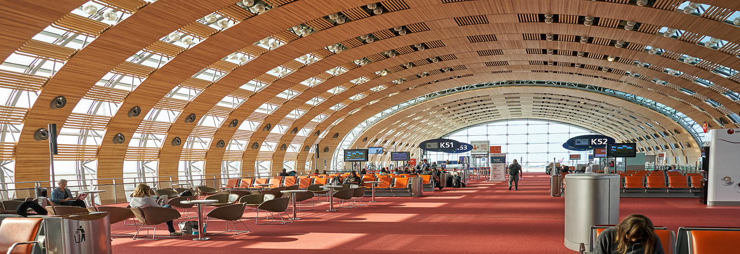 A spacious airport terminal with a curved wooden ceiling, large windows, rows of orange seats, digital gate signs, and passengers waiting near the gates—just steps away from convenient WallyPark Airport Parking.