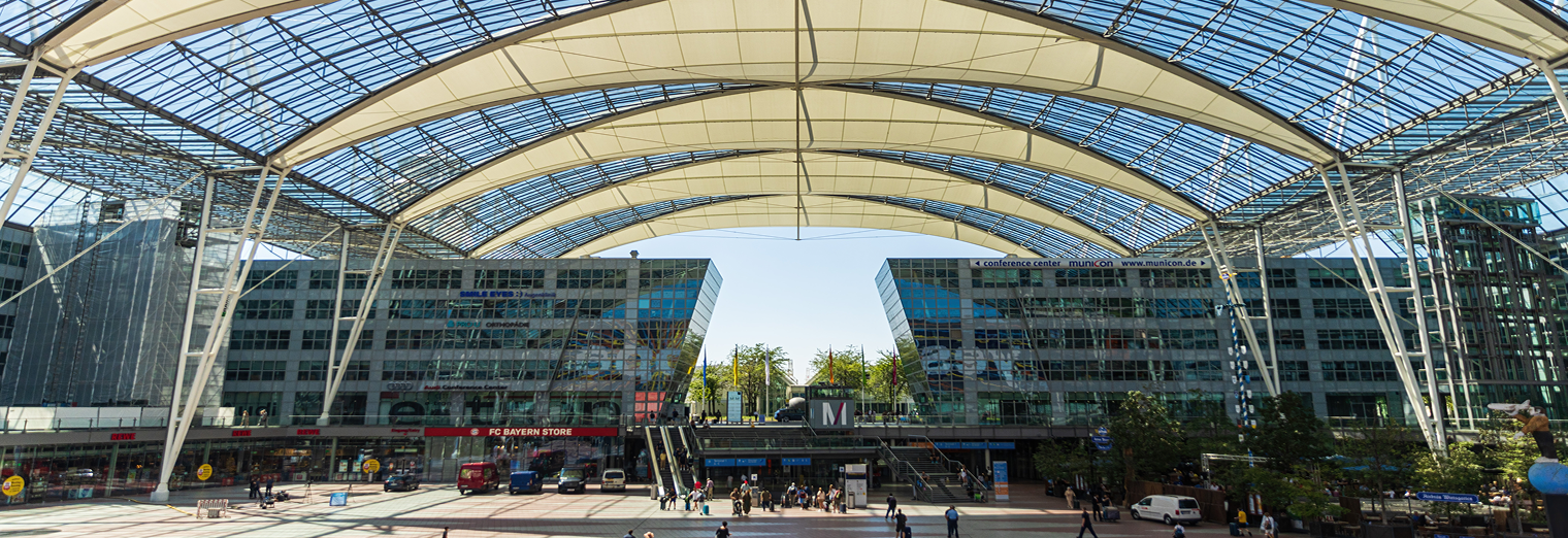 A wide view of Munich Airport’s glass-roofed outdoor plaza, with modern buildings and shops, as people stroll beneath the curved steel and glass canopy—perfect for travelers seeking convenience like WallyPark Airport Parking.