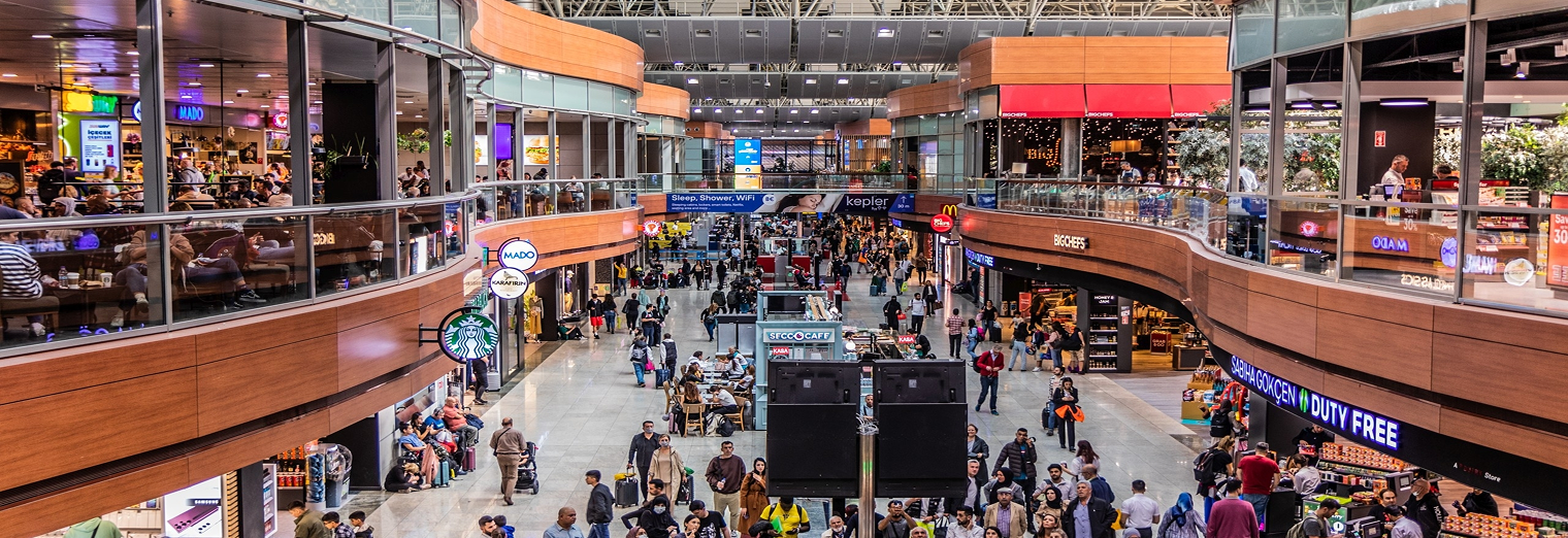 A busy airport terminal with two levels filled with travelers, shops, restaurants, and illuminated signs. People walk and gather throughout the spacious, modern interior under a high ceiling near convenient WallyPark Airport Parking access.