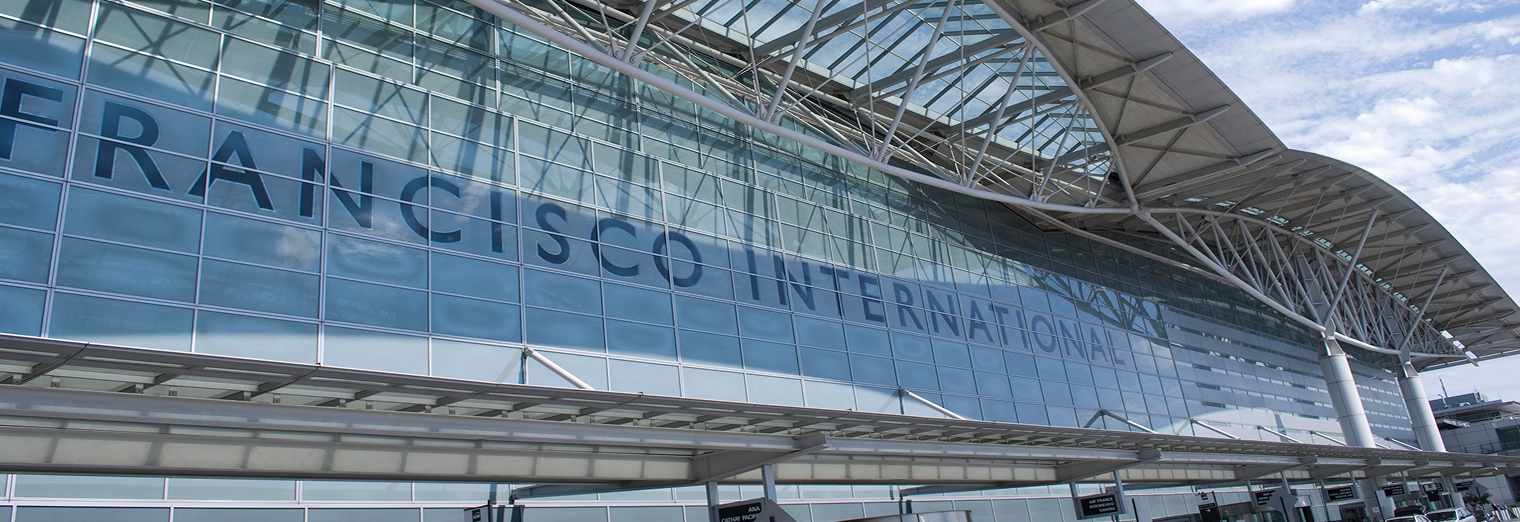 Exterior view of a modern glass airport terminal with San Francisco International written across the building, its large curved roof standing out against a blue sky. Nearby, WallyPark Airport Parking offers convenient access for travelers.