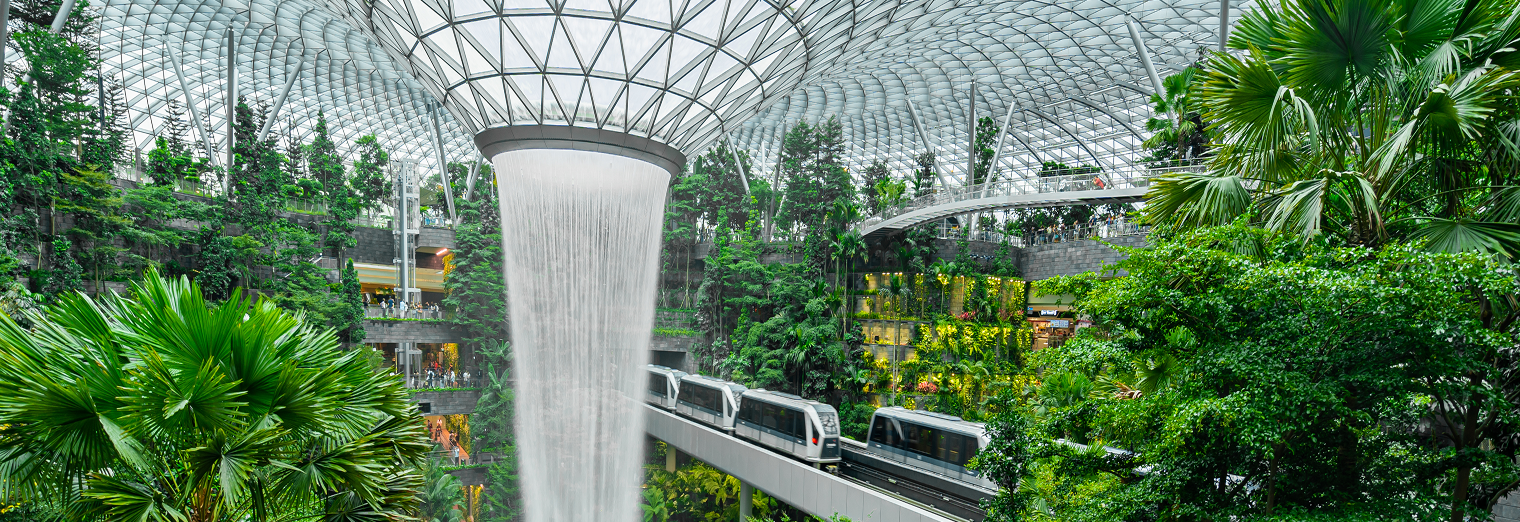 Indoor garden with lush green plants, a glass-domed roof, a large central waterfall, and an elevated train passing through—reminiscent of the tranquility you feel after using WallyPark Airport Parking for stress-free travel.