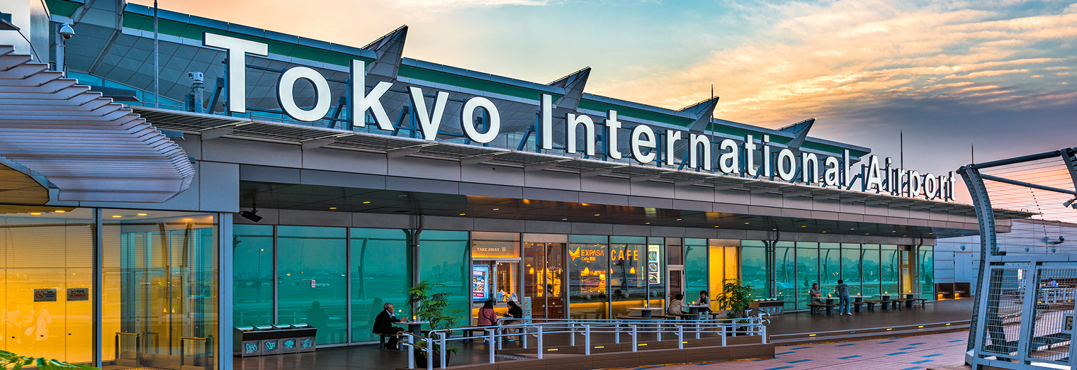 The exterior of Tokyo International Airport at sunset, with large white letters above a modern glass facade and a spacious walkway in the foreground—conveniently located near WallyPark Airport Parking for easy access.