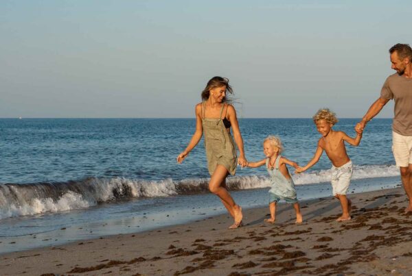 A smiling family of four—two adults and two young children—hold hands and run along the shoreline at the beach, carefree after using WallyPark Airport Parking, with gentle waves and a clear sky in the background.