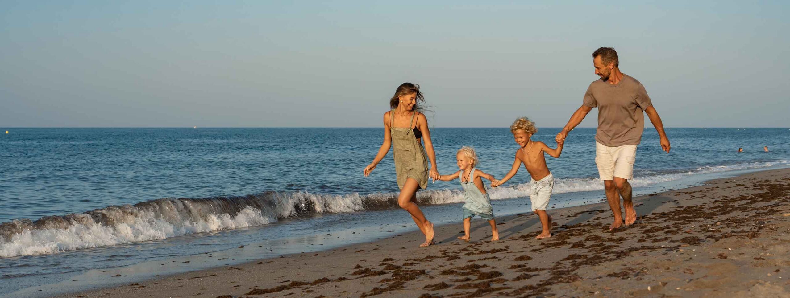 A smiling family of four—two adults and two young children—hold hands and run along the shoreline at the beach, carefree after using WallyPark Airport Parking, with gentle waves and a clear sky in the background.