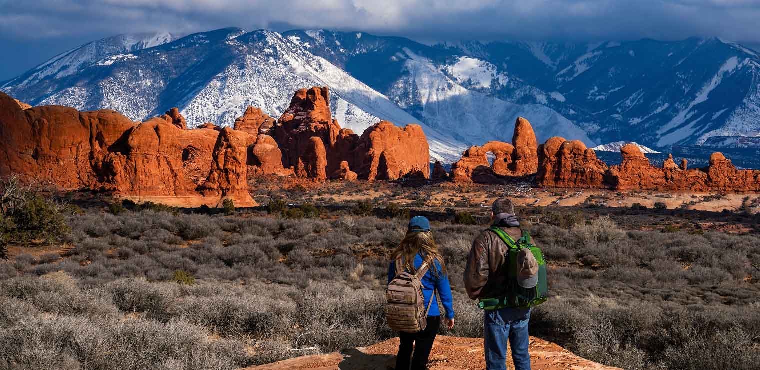 Two hikers with backpacks stand on a rocky path overlooking red rock formations and snow-capped mountains under a dramatic, cloudy sky—reminiscent of the adventures that begin after parking at WallyPark Airport Parking.