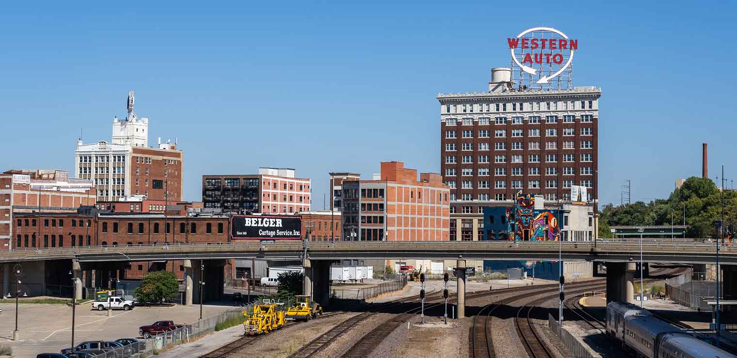 View of downtown Kansas City, featuring railroad tracks, an overpass, brick buildings, the historic Western Auto sign atop a tall building, and clear blue skies—all just a short drive from WallyPark Airport Parking.