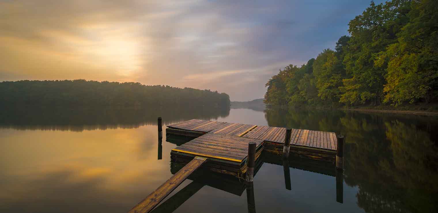 A wooden dock extends over a calm lake surrounded by dense, green trees at sunrise or sunset, with soft, golden light reflecting on the water—a tranquil escape reminiscent of the ease and peace you’ll find with WallyPark Airport Parking.
