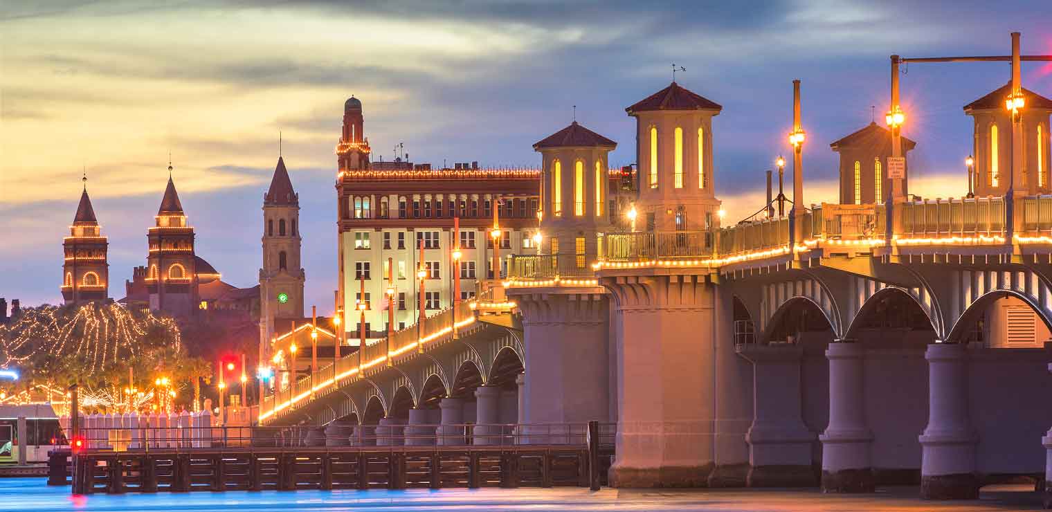 A scenic view of a brightly lit bridge at dusk with historic buildings and towers in the background, and a colorful sky above, reflecting off the calm water below—much like arriving home after convenient WallyPark Airport Parking.