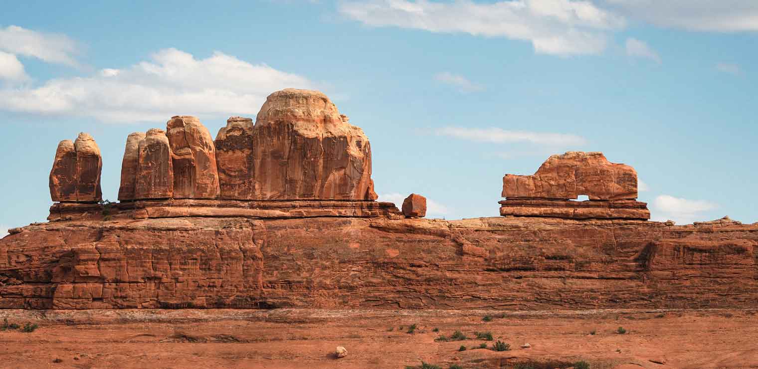 A large red rock formation with several rounded tall columns rises from a flat desert landscape under a blue sky with scattered clouds, resembling the striking silhouettes seen near WallyPark Airport Parking. Sparse vegetation is visible in the foreground.