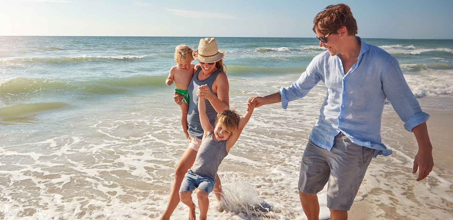 A family of four enjoys a sunny day at the beach. After using WallyPark Airport Parking, the mother holds a child and the father swings another by the arms as they all play at the water’s edge with waves in the background.