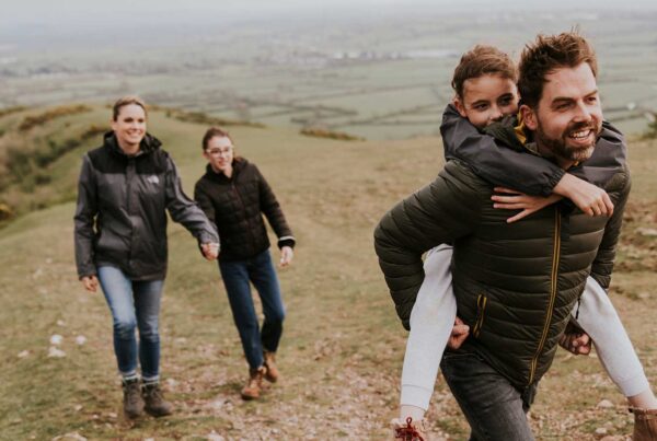 A man gives a young girl a piggyback ride while another adult and child walk behind them, enjoying the outdoors on a grassy hillside with fields in the background—making carefree memories before heading to WallyPark Airport Parking for their next adventure.
