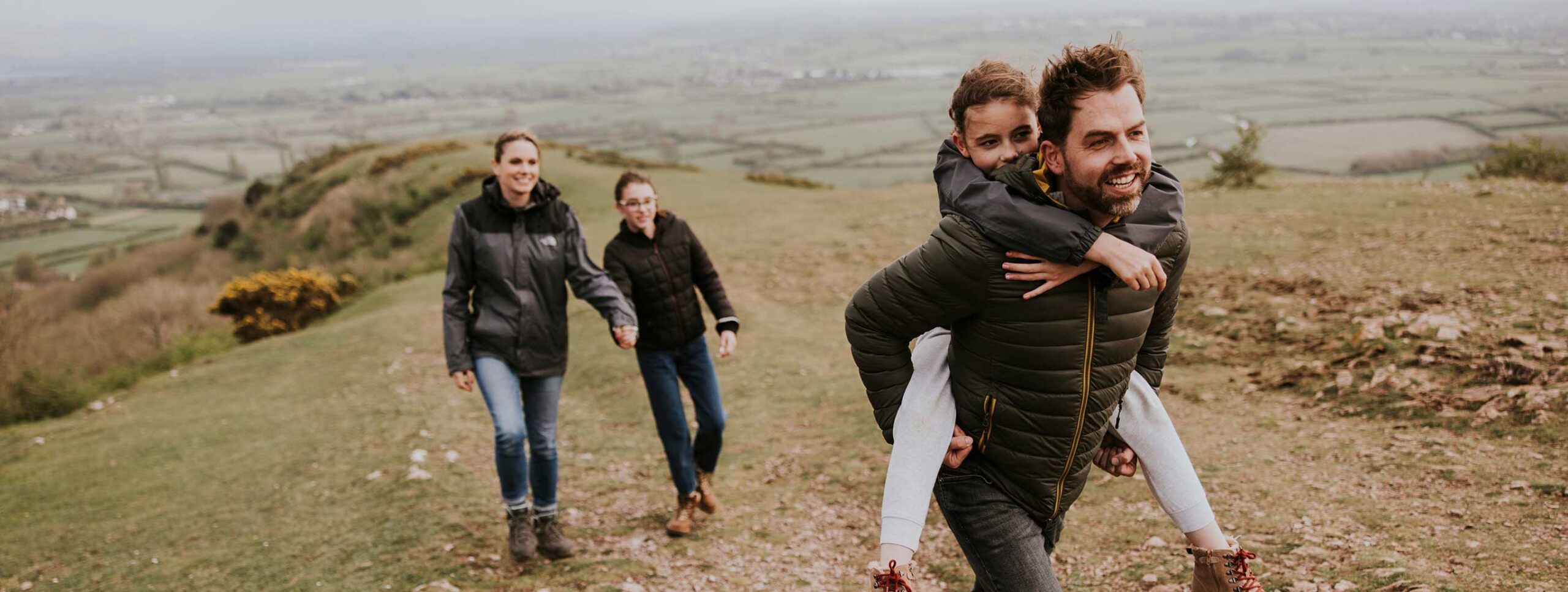 A man gives a young girl a piggyback ride while another adult and child walk behind them, enjoying the outdoors on a grassy hillside with fields in the background—making carefree memories before heading to WallyPark Airport Parking for their next adventure.