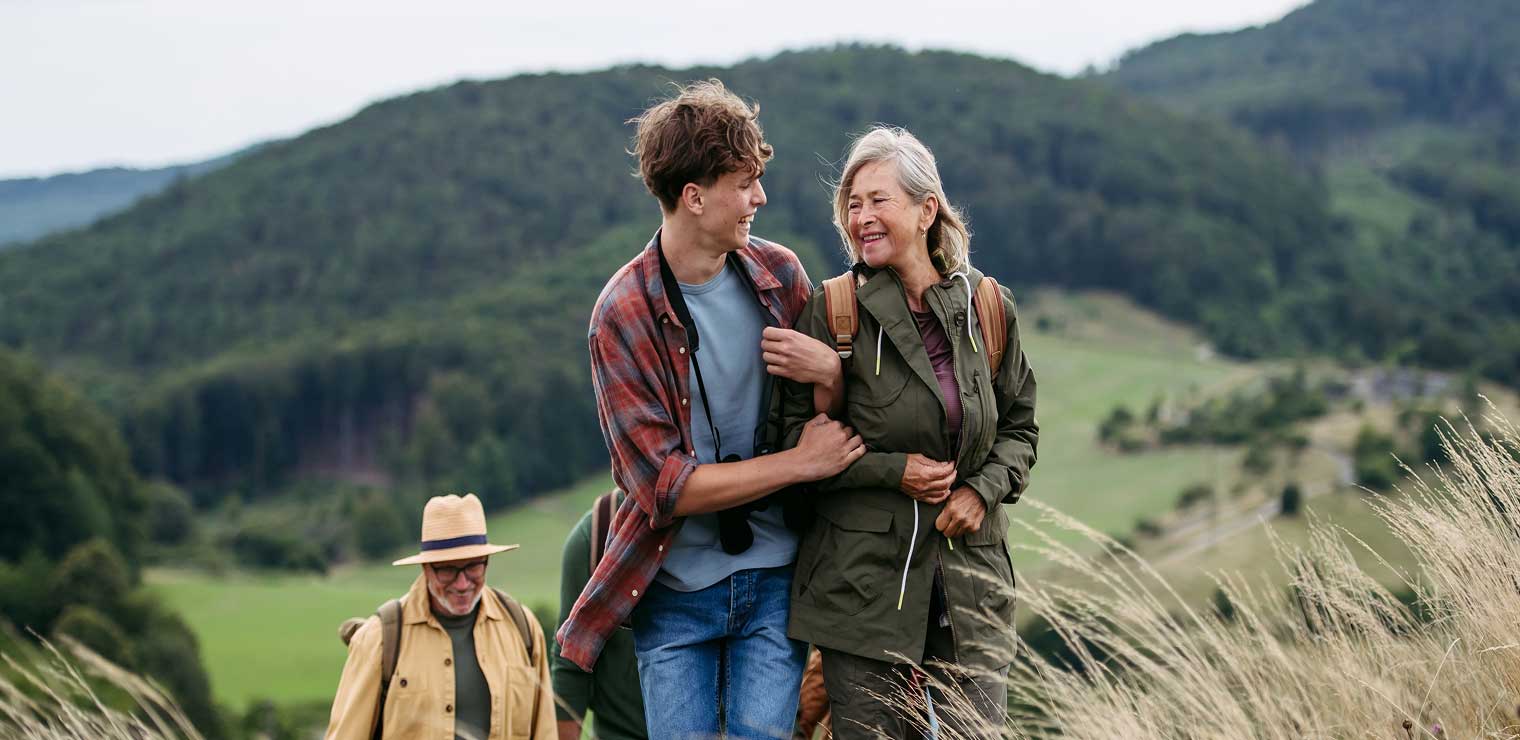 Three people hike in grassy hills; a young man and an older woman smile and walk arm in arm, while an older man in a hat follows behind. Reminiscent of the carefree feeling after securing your car at WallyPark Airport Parking, forested hills and fields stretch out in the background.