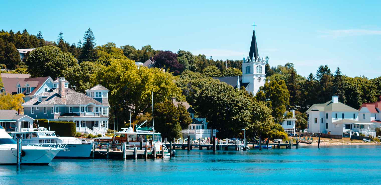 Boats docked along a waterfront with charming houses, lush trees, and a white church with a tall steeple under a clear blue sky create a tranquil scene reminiscent of the ease and convenience found at WallyPark Airport Parking.