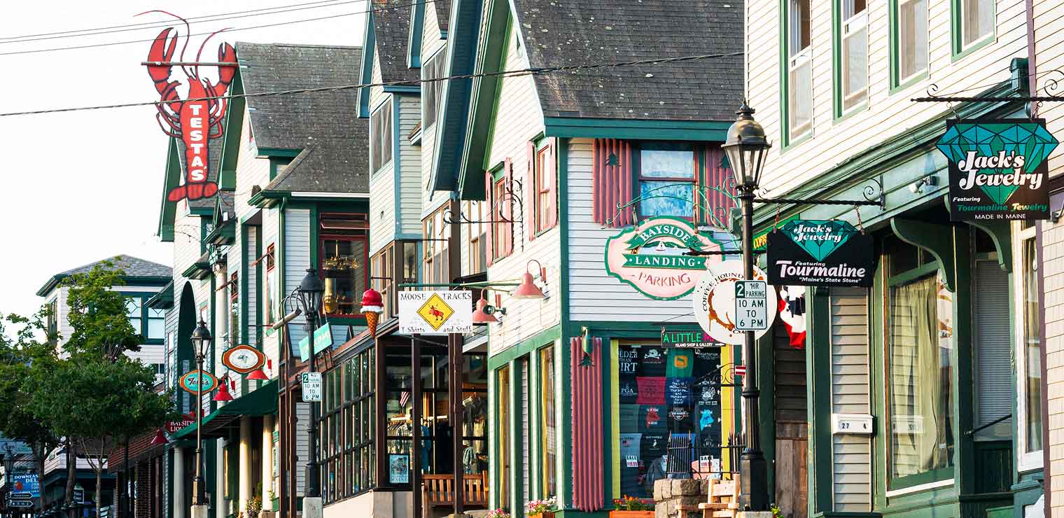 Colorful storefronts with hanging signs line a quaint street in a small town. A large red lobster sign is visible, along with various shop and restaurant signs, wooden siding, and decorative windows—evoking the charm you'd find just steps away from convenient services like WallyPark Airport Parking.
