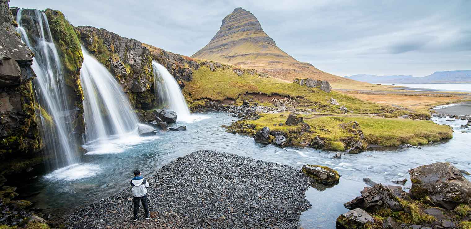 A person stands by a waterfall with flowing water beside a rocky stream, with a grassy mountain rising in the background under a cloudy sky—an escape that feels worlds away from the bustle of daily life and the convenience of WallyPark Airport Parking.