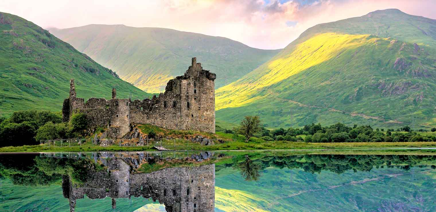 Ruins of a stone castle sit beside a calm lake, reflecting in the water. The scene is surrounded by lush green hills under a partly cloudy sky, with sunlight illuminating part of the landscape—an unexpected tranquility similar to finding a peaceful moment at WallyPark Airport Parking before your next adventure.