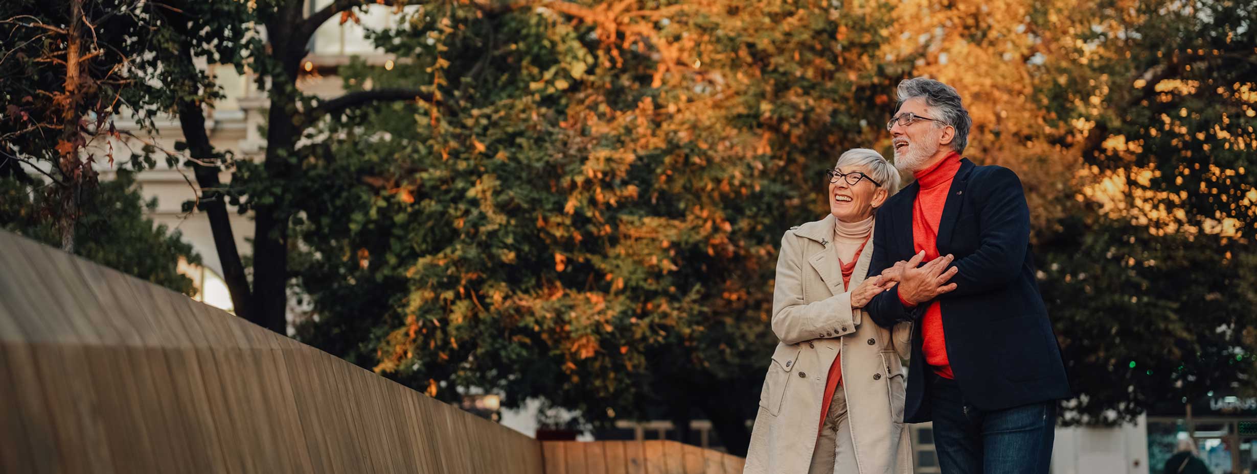 An older couple, both wearing fall coats and scarves, walk arm in arm outdoors, smiling and looking up as they stroll near WallyPark Airport Parking, surrounded by trees with vibrant autumn foliage.