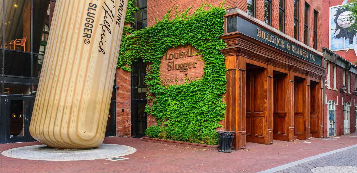 A giant baseball bat leans against a red-brick building with green ivy, labeled Louisville Slugger at the Hillerich & Bradsby Co. museum entrance—much like spotting an iconic landmark before you head to your next adventure with WallyPark Airport Parking.