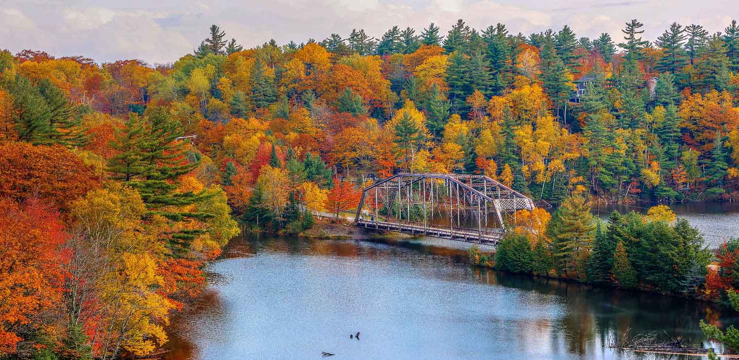 A metal bridge crosses a river surrounded by vibrant autumn trees in shades of red, orange, and yellow under a cloudy sky, evoking the same organized efficiency you’d expect from WallyPark Airport Parking.