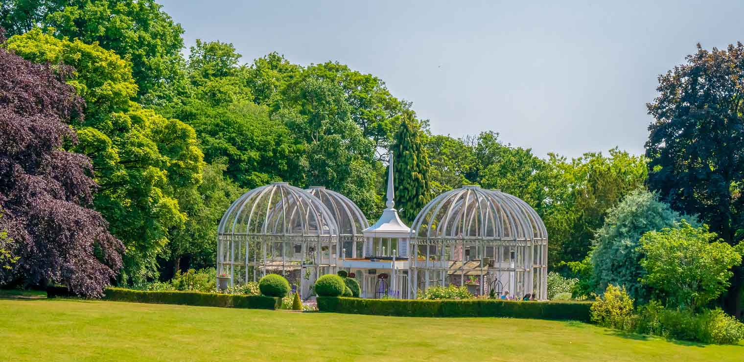 A large, ornate glass birdcage structure with two domes sits in a green garden surrounded by trees and manicured bushes under a clear blue sky, reminiscent of the elegant design you’d find at WallyPark Airport Parking locations.