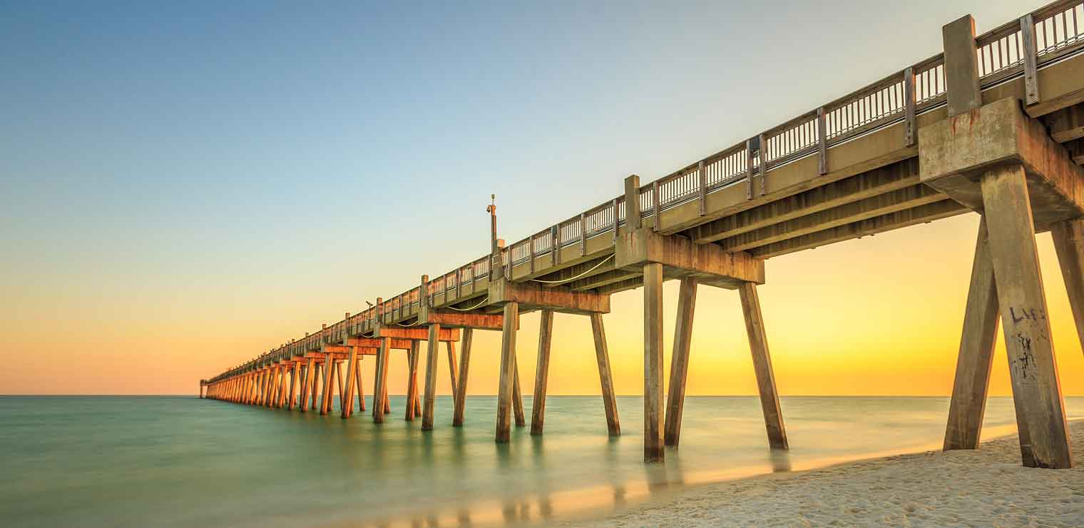 A long concrete pier stretches over calm, green water toward the horizon at sunset, with a clear sky and sandy beach in the foreground—offering a tranquil escape reminiscent of arriving at your destination after the convenience of WallyPark Airport Parking.