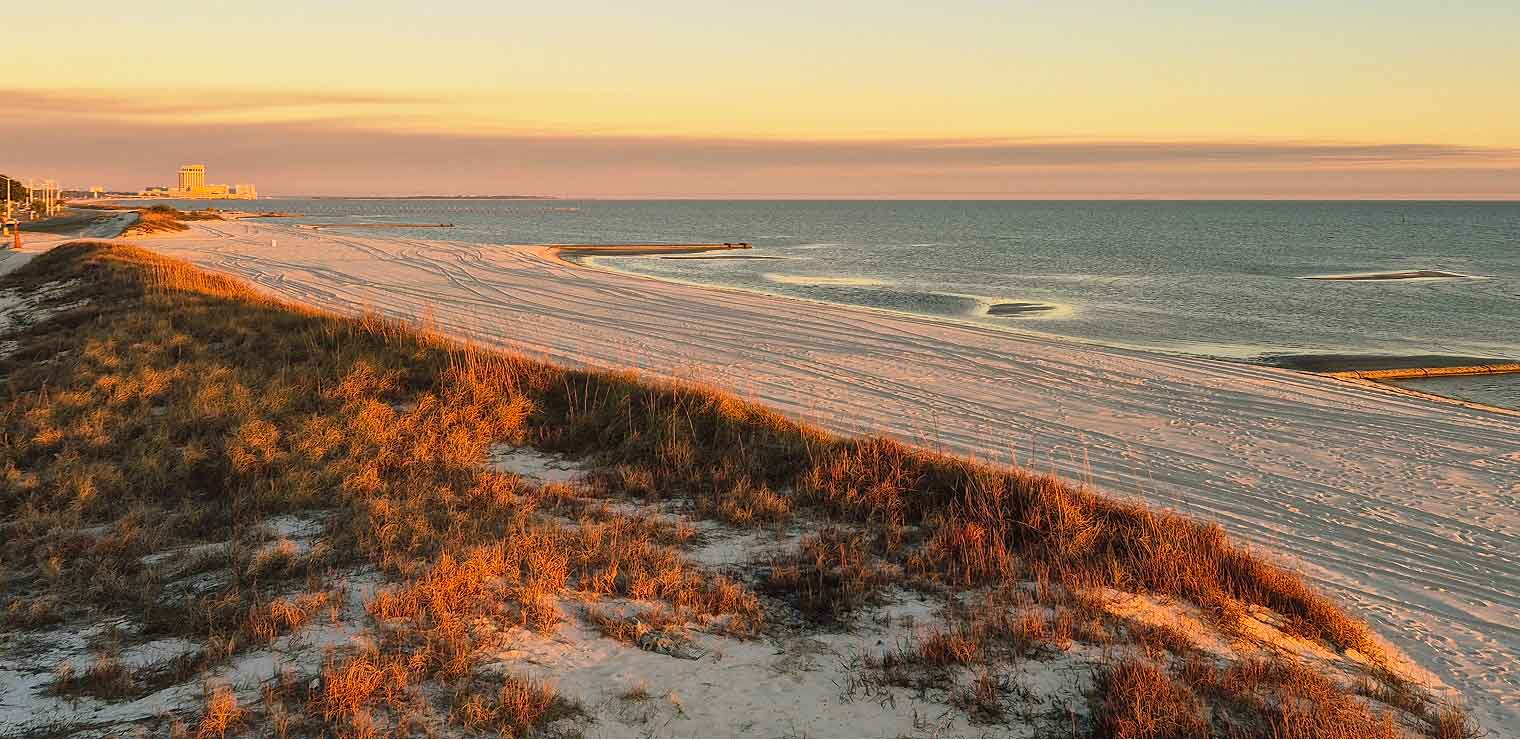A sandy beach at sunset with gentle waves, grassy dunes in the foreground, and buildings visible in the distance along the shoreline—just a short drive from WallyPark Airport Parking.